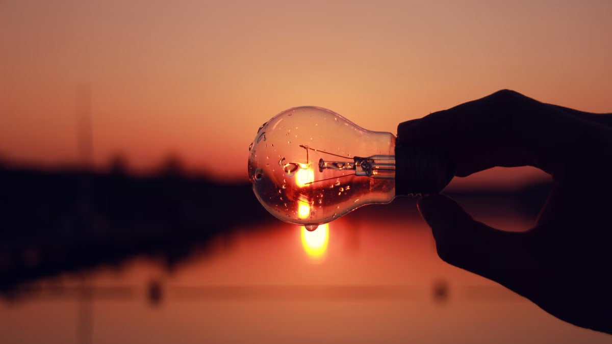 Silhouette of a hand holding a light bulb against a stunning sunset over a lake, blending creativity and nature.